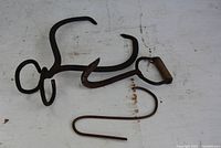 Metal hand grappling tool, ice thongs, thick metal hanger lying on white surface showing rust and wear