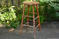Full view of wooden bar height stool outdoors against greenery, showing round top and four legs with cross supports.