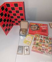 The full lot viewed on a table showing all included items: folding checkered game board, Television Stars puzzle box, Heirloom Seeds puzzle box, playing cards, flash cards.