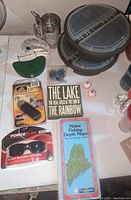 Fishing gear assortment showing tackle boxes, fishing bobbers, books, a fish bite alarm, and other accessories on a white table