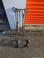 Fireplace tools set on stand including poker, tongs, brush, shovel and two iron andirons placed on the ground in front.
