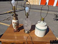 Three table lamps displayed outdoors on wooden surface; includes ceramic floral design lamp, crystal cut glass lamp, and white ceramic jar-shaped lamp