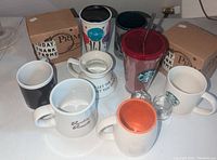 Eight assorted coffee mugs and tumblers displayed on a surface including Starbucks, Dunkin Donuts, Primitive by Kathy, and Elvis themes