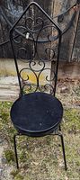 Black metal Mid-Century Modern style chair with decorative swirl backrest and round seat photographed outdoors against a wooden fence.