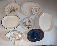 Top down view of seven plates and dishes arranged on a white surface including Goebel fine china plate, bird illustration plate, and various oval dishes