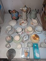 Photo showing a collection of various teapots, cups, saucers, creamers, and small accessories arranged on white surface against plain background.