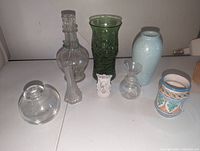 Seven assorted vases and containers arranged on a white surface, showing clear glass, green glass, small decorative ceramic, and textured blue vase.
