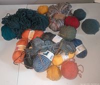 Mixed yarn skeins and balls on a white surface, mainly wool, some new with labels, some unwrapped in colors like teal, orange, mustard, red, green, grey, blue.