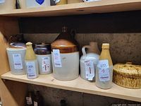 Shelf display showing large two-tone brown and beige jug, tan ceramic bottle with cork stopper, and other assorted stoneware crocks and jugs with visible auction lot labels.
