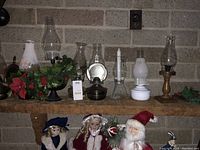 Shelf display of 8 assorted vintage non-electric oil lamps along with decorative greenery and dolls seen below the shelf.