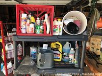 Lot overview showing plastic shelving with automotive liquids and containers including red crate with sprays, jugs, and funnel bucket.
