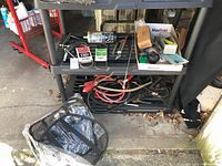 Two garage shelves with automotive tools and accessories including jumper cables, siphons, and various small items organized loosely. The items are stored on black plastic shelving units with some leaves scattered on the floor.