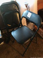 Set of stacked black cushioned folding chairs seen from the front and side, showing wear, some peeling and scrapes on cushions and frames.