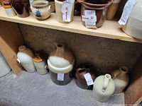 Photo showing six ceramic crocks or jugs on a bottom shelf, varying in size from small to medium, all with handles and mostly beige and brown glaze.