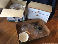 Image showing vintage wooden bread box, electric crock pot box, various glass lids, and white ceramic bowl placed on cardboard and wooden floor.