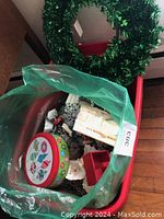 Photo shows a green shamrock wreath hanging on a red chair. Inside a green plastic bag on the chair are several holiday decor items: a round Christmas tin, a small white artificial Christmas tree, a red box, and a white bag with Christmas text design.