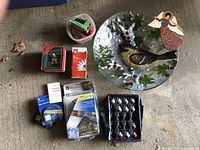 Overview of lot showing all items on concrete floor outdoors. Includes glass platter with festive cardinal and holly, boxed Christmas lights, small figurine ornament, light rope in packaging, assorted bulbs, and angel cut-out.