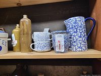 Shelf with blue and white spongeware soup bowls stacked, a blue and white spongeware pitcher on the right, and stoneware bottles on the left.