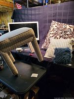 View showing ottoman on black foldable table, front side with angled legs, fabric with diamond pattern, and three throw pillows behind it on black backdrop.