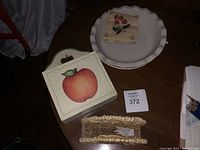 Photo showing cream ceramic plate, wooden box with apple painting, and woven napkin holder