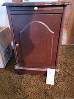 Wooden magazine rack showing single door with curved panel and metallic knob on carpet.