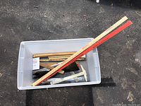 Plastic bin containing multiple wooden, plastic, and metal rulers including yardsticks, with some printed logos and measurement scales visible.