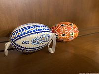 Two hand-painted decorative eggs placed on a wooden surface, one blue and white with yellow accents and the other red with beige and white designs