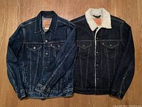 Two Levi's denim jackets side by side on wooden floor, one with sherpa collar and lining, the other classic denim jacket.