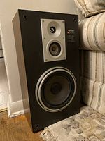 Photo showing one Technics SB-L51 speaker standing upright near a couch, displaying front drivers and wood finish sides.