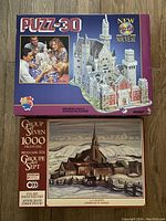 Two unopened puzzle boxes stacked: a purple Puzz-3D Bavarian Castle 3D puzzle box and below it a Group of Seven 1000-piece puzzle box featuring an artwork of a church scene.