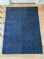 Black door mat with patterned square floral tiles and border placed on wooden floor in front of a closed door.