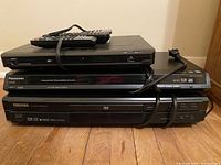 Three black DVD players stacked with visible front panels and display screens, each with a compatible remote and attached power cords laid on wooden floor.