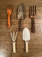 Five vintage gardening tools on wooden floor: three metal and wood tools showing rust (two mini rakes and one trowel) and two cream plastic-handled tools (one mini rake, one trowel).