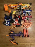 Top-down photo of entire collection showing Vulcan blaster, Big Bad Bow, two smaller blasters, two dart vests, drum magazine, and multiple darts and foam tubes on wooden floor.