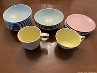 Photo showing stacks of blue plates and bowls, pink small plates, and two yellow handled cups on a wooden surface.