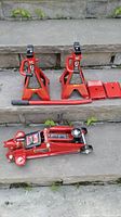 Photo showing red 2 ton hydraulic floor jack, two red 2 ton jack stands, jack handle, and two red metal base plates on concrete steps outdoors.