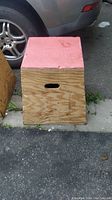 Front and side view of the wooden plyometric box showing red padded top, natural wood ply finish, and hand grip cutout on side.