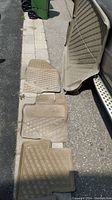 Six beige rubber car mats and trunk mats from Volvo XC70 laid out for display on an outdoor pavement surface. Mats show dirt, wear use marks and traction patterns.