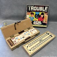 Two board games side by side; open cribbage pegless board game in beige case showing numbered tiles and scoring board; Trouble game box behind.