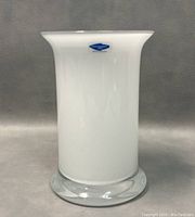 Front view of a white glass vase with a clear glass base and a blue label sticker near the top, sitting on a gray background.
