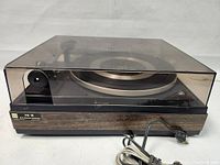 Side-front view of the Dual CS 16 turntable with closed plexiglass dust cover and visible wood grain front panel, power cord coiled in front.