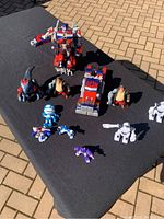 Two large Optimus Prime Transformers, one in robot form and one in vehicle form missing a wheel, with various smaller toys including robotic animals and humanoid figures arranged on a black cloth outdoors.