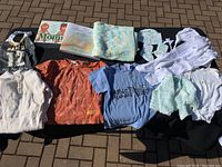 Overview of all items in the lot showing books, bib, various baby shirts, knitted blanket, bonnet, booties, and onesies laid out on the ground.