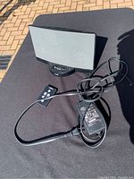 Bose speaker with front grille, remote control, and power adapter seen on outdoor table with sunlight.