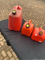Three red fuel cans on black cloth; one large 20 litre can and two smaller 5 litre cans, one missing top of cap