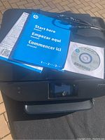 HP OfficeJet 5740 printer unit with manuals and installation CD on top, showing front and top views of the device with paper trays and display screen visible.