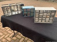 Three hardware storage containers with many small clear drawers labeled and filled with assorted screws, washers, and pins, placed on a black tablecloth outdoors.