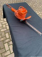 Full view of the orange Black & Decker electric hedge trimmer placed on a black surface showing the blade, handle, and coiled power cord.