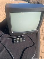 Small black CRT television and matching black remote control displayed on a dark surface outdoors, showing TV front with screen and attached power cable.