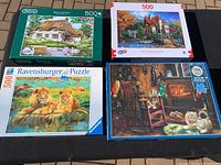 Four 500-piece puzzle boxes arranged on ground. Images include Swan Cottage, an autumn lake house, cozy fireplace with cats, and lion family.